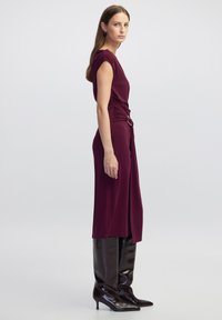 Burgunderfarbenes Midi-Kleid mit einem drapierten Seitendesign und silberner Hardware, kombiniert mit kniehohen, glänzenden schwarzen Stiefeln mit spitzer Zehe.