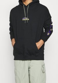 Felpa nera con cappuccio, tasca centrale, e design grafico sul petto. Presenta dettagli viola e oro sulle maniche. Materiale in cotone.