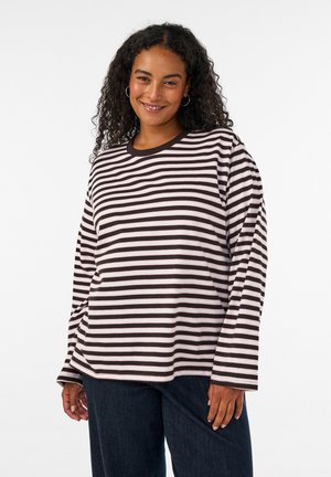 Femme souriante aux cheveux bouclés, portant une chemise rayée noir et blanc à manches longues et un jean foncé, debout devant un fond blanc uni.
