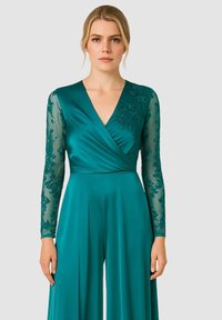 Tuta color teal con maniche lunghe in tessuto trasparente, adornata con ricami floreali intricati e design a scollo a v, realizzata in tessuto liscio.