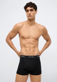 Homem musculoso a usar boxers pretos da Calvin Klein, em pé com as mãos nos quadris, olhando ligeiramente de lado contra um fundo simples.