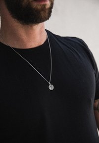 Strieborný retiazkový náhrdelník s kruhovým príveskom so zložitým dizajnom, nosený cez čierne tričko. Hladký kovový povrch a minimalistický štýl.