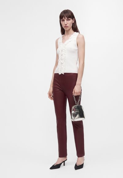Femme portant un haut blanc sans manches à volants et un pantalon rouge foncé ajusté, tenant un petit sac à main argenté avec une bandoulière chaîne.