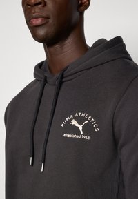 Mørkegrå hoodie med kængurulomme, der har et hvidt broderet Puma Athletics-logo og snore med metalspidser.