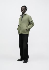 Olive green, lightweight jacket with a collar, zippered pockets, and a relaxed fit, paired with black straight-leg pants and black shoes.