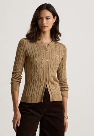 Kvinde med mørkt hår iført en beige kabelstrikket cardigan med knapper og mørke bukser mod en ensfarvet lys baggrund.