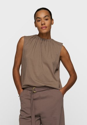 Frau mit den Händen in den Taschen, die ein ärmelloses, taupefarbenes Oberteil mit gerafftem Ausschnitt und passende Gürtelhose vor schlichtem Hintergrund trägt.