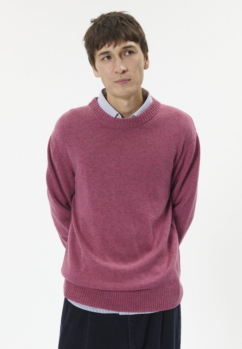 Fuksziaszínű kötött pulóver bordázott kerek nyakkal és mandzsettával, csíkos, galléros ing fölé viselve. A szabás laza, sima textúrájú.