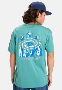 Camiseta de manga corta en color aguamarina con un diseño gráfico azul de montañas y elementos abstractos, con el texto “SOUND WAVES” en la parte inferior.