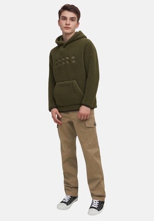 Sudadera de felpa verde olivo con bolsillo frontal y detalles del logo, combinada con pantalones cargo beige y zapatillas negras con suelas blancas.