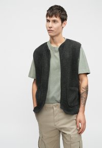 Juodas vilnonis liemenė su šoninėmis kišenėmis, užtrauktuko uždarymu, dėvima virš šviesiai žalio marškinėlių. Modelis vilki šviesiai rudas krovinių kelnes. Paprastas, kasdieniškas dizainas.