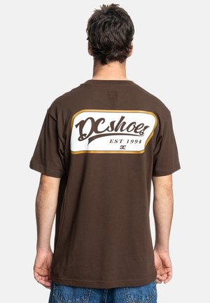Brown short-sleeve t-shirt featuring a large white and gold rectangular logo on the back with the text "DC Shoes EST 1994."