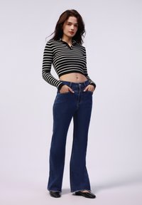 Zwart-wit gestreepte cropped sweater met lange mouwen, gecombineerd met donkerblauwe flared jeans. Model staat met de handen in de zakken.