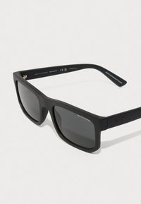 Matē melni saulesbrilles ar tumši pelēkiem stikliem, taisnstūra forma, un platais kātu dizains ar izcelto "Armani Exchange" logotipu.
