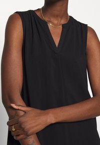 Person med ärmlös svart topp, guldkedja, guldring, med korsade armar och synliga tatueringar på underarmarna mot vit bakgrund.