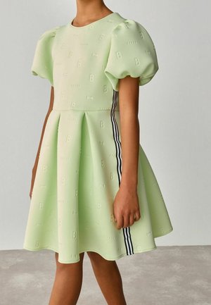 Enfant portant une robe vert pâle à manches bouffantes avec un motif de lettres en relief et des bandes latérales noir et blanc, debout sur un sol gris.