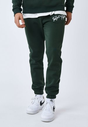 Pantalons de survêtement vert foncé avec taille à cordon et poignets côtelés. Présente une broderie blanche "Project" sur la cuisse gauche. Associés à des baskets blanches.