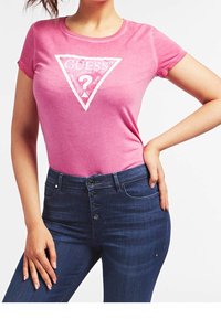 Lyserød bomulds t-shirt med korte ærmer, der har et hvidt trekantet logo med "GUESS" og et spørgsmålstegn i midten. Mørk denim jeans med knapper.