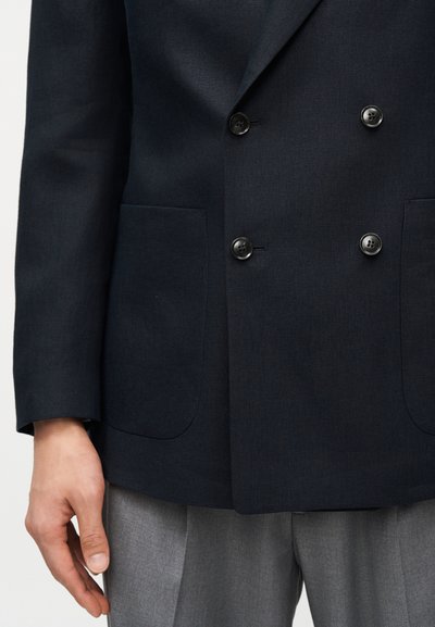 Άνδρας που φοράει σκούρο μπλε διπλομπλεϊζερ με τέσσερα κουμπιά και δύο εξωτερικές τσέπες πάνω από ανοιχτό γκρι παντελόνι.