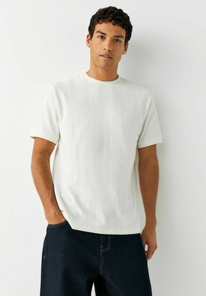 Göndör hajú férfi, fehér texturált rövid ujjú inget és sötét farmert viselve, fehér, egyszínű háttér előtt áll.