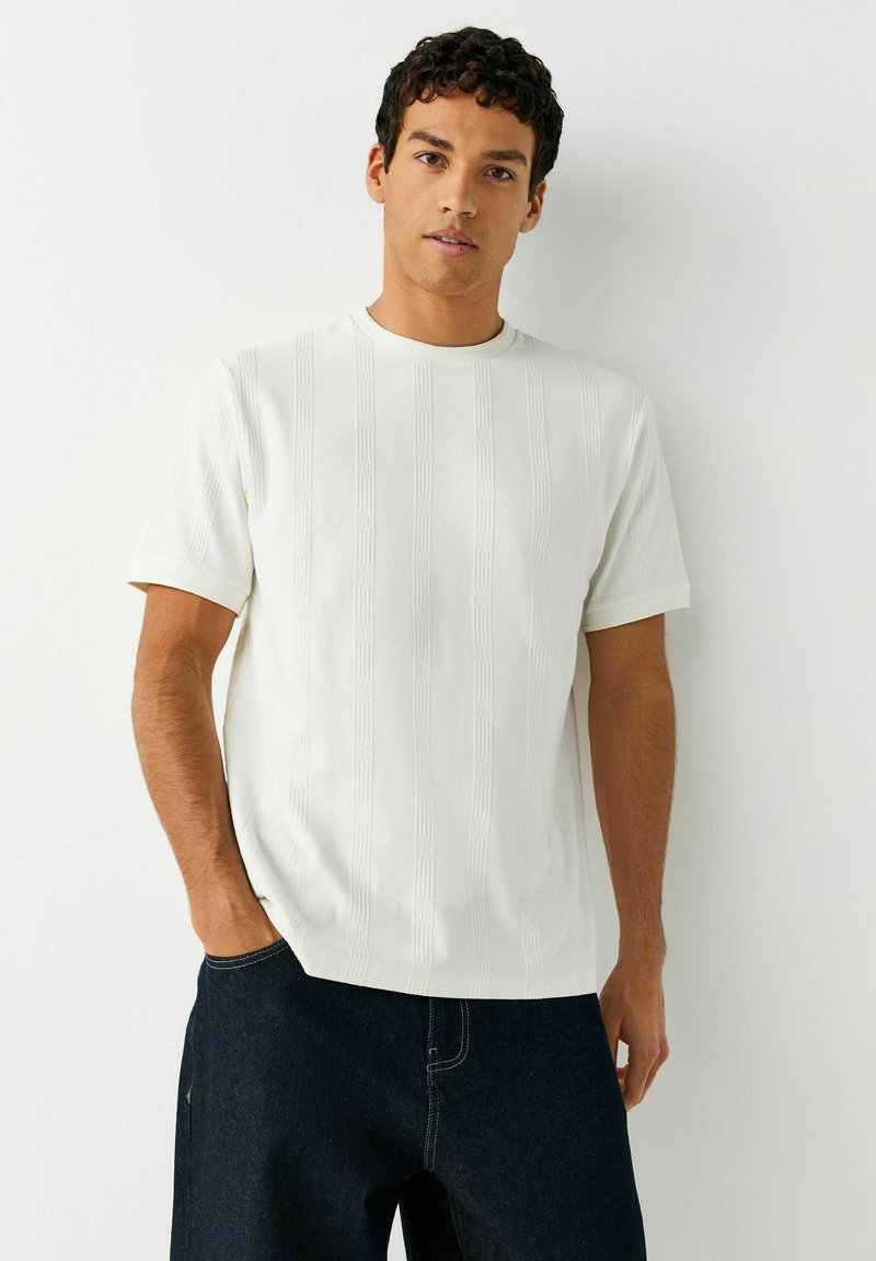 Uomo con capelli ricci che indossa una camicia bianca a maniche corte con tessuto strutturato e jeans scuri, in piedi davanti a uno sfondo bianco semplice.