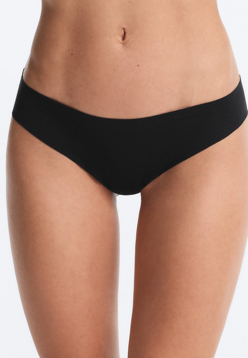 Femme portant des sous-vêtements noirs sans coutures de style bikini, montrant l'abdomen et le haut des cuisses sur un fond clair uni.