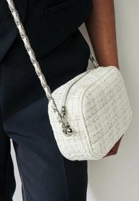 Witte tweed schoudertas met een rechthoekige vorm, zilveren hardware, ritssluiting en een gedraaide kettingband. Geprofileerde oppervlakte met subtiele patronen.