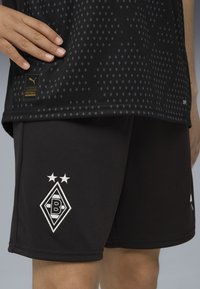 Hand auf einem schwarzen Fußballtrikot mit Rautenmuster; schwarze Shorts zeigen ein weißes, diamantförmiges Logo mit zwei Sternen darüber.