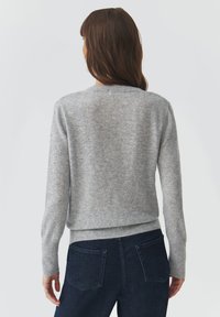 Grå strikket sweater med rund halsudskæring, ribbede manchetter og kant, med en blød tekstur. Sammen med mørke denim bukser, set fra bagsiden.