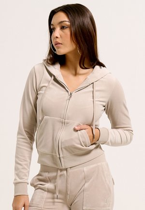 Jonge vrouw met lang bruin haar draagt een beige hoodie met rits en bijpassende broek, kijkt naar de zijkant met een hand in haar zak.