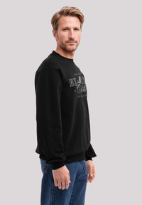 Schwarzer Sweatshirt mit Rundhalsausschnitt, gerippten Bündchen und einem grafischen Druck in weißem Text. Tragen mit blauen Jeans.