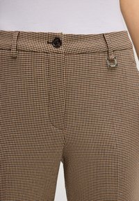 Pantalons à motif pied-de-poule marron avec une poche latérale, fermeture à bouton et accent logo en métal. Tissu texturé avec une coupe ajustée.