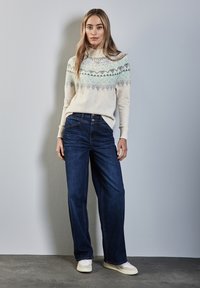 Licht crèmekleurig sweater met geometrische blauwe patronen, gecombineerd met donkerblauwe wijde jeans en witte sneakers. Effen achtergrond met zachte belichting.