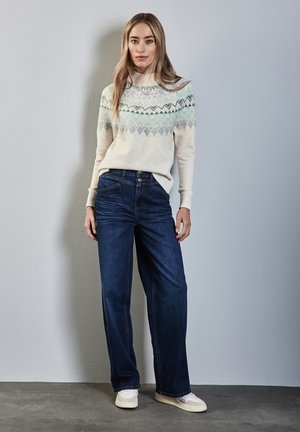 Pull léger en crème avec des motifs géométriques bleus, associé à un jean large bleu foncé et des baskets blanches. Fond uni avec un éclairage doux.