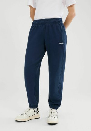 Persona che indossa pantaloni della tuta Ellesse blu navy e scarpe da ginnastica bianche, in piedi con le mani dietro la schiena contro uno sfondo semplice.