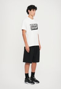 Balta T-kreklu ar melnu "Versace Jeans Couture" logotipu, apvienojot ar melniem šortiem un melniem apaviem. Apģērbs ir ar brīvu piegriezumu.