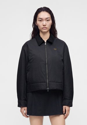 Femme aux longs cheveux foncés porte une veste noire zippée avec col et une courte jupe noire, debout devant un fond clair uni.