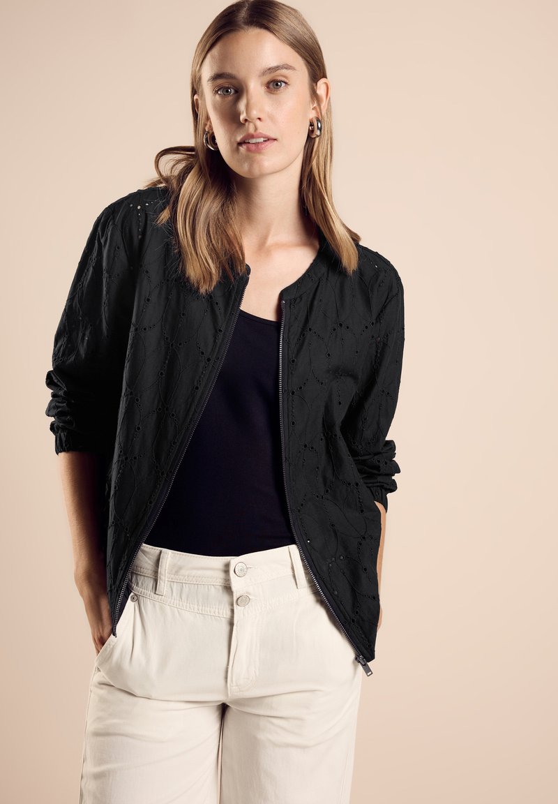 Veste bomber noire avec un motif texturé, des poignets côtelés et une fermeture éclair, associée à un haut noir et un pantalon de couleur claire.