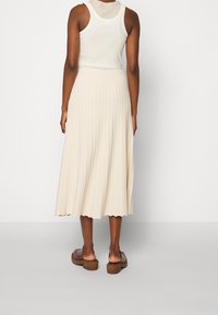 Beige plisat midi-skjørt med en teksturert finish, kombinert med en hvit ermeløs topp og brune hælsandaler. Skjortekanten har bølget detalj.