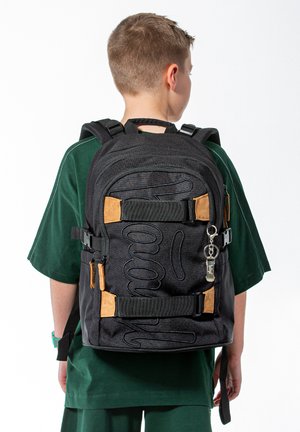 Junge trägt dunkelgrünes Hemd und großen schwarzen Rucksack mit braunen Akzenten, steht mit dem Rücken zum weißen Hintergrund.