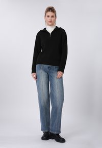 Pull-over noir en tricot avec col à demi-fermeture éclair, associé à un jean large bleu clair et des bottines noires. Textures lisses, coupe décontractée.