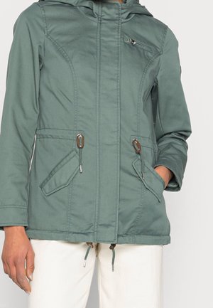 Personne portant une veste à capuche verte avec une poche zippée sur la poitrine, des poches à rabat, des cordons de serrage à la taille et un pantalon blanc.