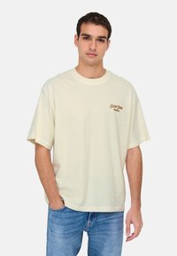 Lyscremet kortærmet t-shirt lavet af bomuld. Har et lille brunt logo på venstre bryst og en afslappet pasform. Matchet med blå jeans.
