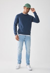 Man in groene pet, marineblauwe sweatshirt, lichtblauwe jeans en witte sneakers staat tegen een witte achtergrond, terwijl hij met zijn rechterhand zijn pet aanpast.