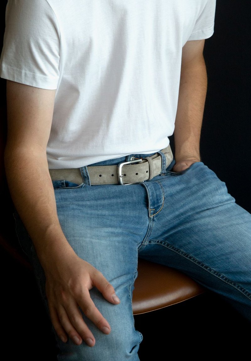 Lichtgrijze suède riem met een zilveren metalen gesp, gedragen over een blauwe spijkerbroek, gecombineerd met een wit T-shirt, zittend op een bruine stoel.