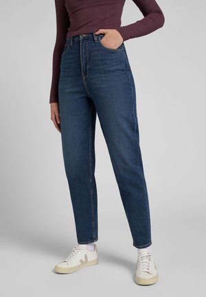 Osoba ubrana w ciemnoniebieskie dżinsy z wysokim stanem, białe trampki i bordową bluzkę z długim rękawem, z jedną ręką w kieszeni, stojąca na szarym tle.