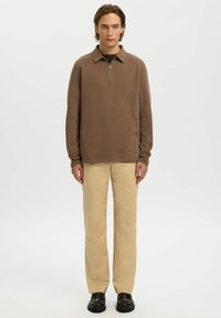 Brun långärmad pullover med dragkedja vid kragen och texturerat tyg, kombinerad med beige byxor med raka ben och svarta skor.