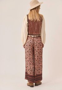 Mujer con sombrero beige, blusa crema de mangas largas, chaleco de terciopelo marrón, pantalones anchos con estampado floral y botas marrones, de pie de espaldas.