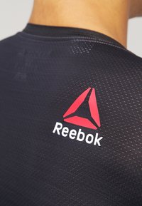 Černé sportovní tričko s texturovaným vzorem, s červeným a černým logem Reebok umístěným nad výstřihem na zadní straně.