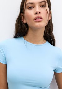 Világoskék, testhezálló, rövid ujjú felső, kerek nyakkivágással. Simább textúrája és varrás nélküli kialakítása kiemeli a karcsú megjelenést.